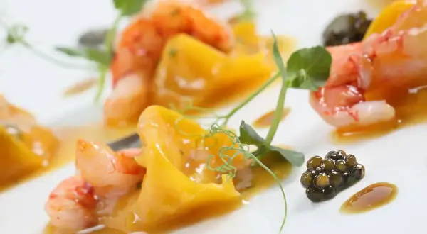 Gamberi succulenti e caviale nero su un letto di pasta con pesce, guarnito con germogli verdi e una salsa dorata.