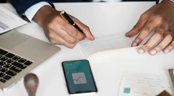 Uomo d'affari scrive su un libretto, con laptop e smartphone che mostrano grafici finanziari. Un esempio di come la categoria protetta art. 1 possa operare nel mondo del lavoro.