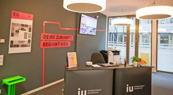 Hier ist die Rezeption der IU Internationale Hochschule. Ein Bildschirm zeigt Bilder, und ein Plan weist den Weg. Was ist die IU? Eine Hochschule, die deine Zukunft beginnt.