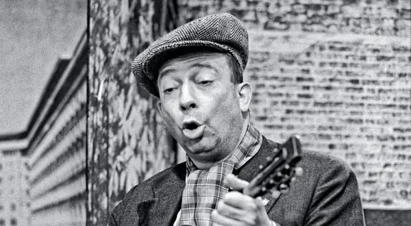 Stanisław Bard Starej Warszawy, w czapce i z gitarą, śpiewa na tle ceglanej ściany.