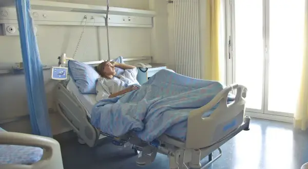 Młoda kobieta leży w szpitalnym łóżku, otulona niebieskim kocem. Jej hospitalizacja przebiega spokojnie, a przez okno wpada jasne światło.