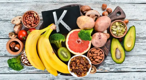 Frutta e verdura che mostrano dove si trova la vitamina K2: banane, broccoli, avocado, pomodori, uva passa, mandorle, semi di zucca, patate, funghi, nocciole.