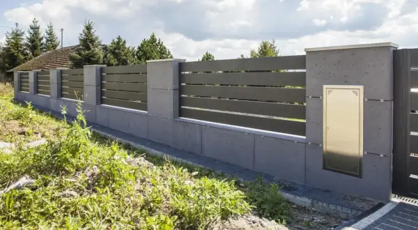 Nowoczesne ogrodzenie z betonowych słupków i ciemnych paneli, wyznaczające nieprzekraczalną linię zabudowy. Złota skrzynka na listy wmurowana w słupek.