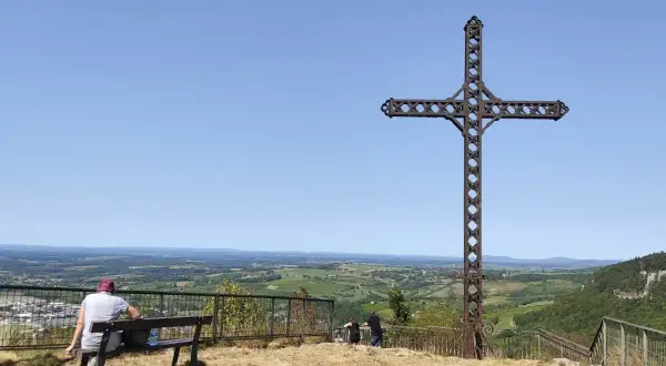 La croix du Dan domine un paysage verdoyant. Deux personnes profitent de la vue depuis un banc.