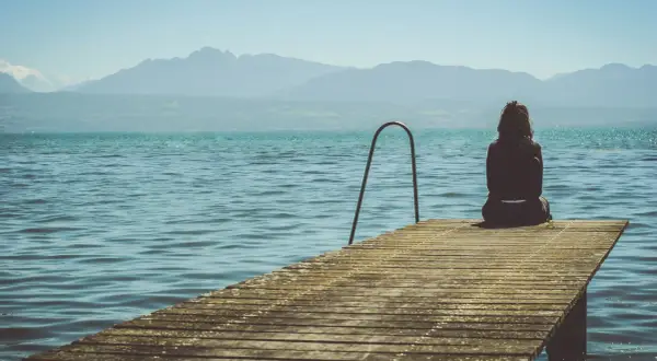 Mujer sentada en un muelle, contemplando el lago y las montañas. Pensamientos de frases de preocuparse por alguien flotan en el aire.