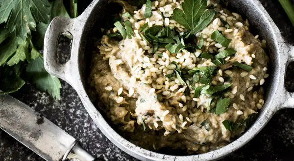 Cremiges Baba Ganoush, garniert mit Sesam und Petersilie, in einem rustikalen Topf. Daneben ein Messer und frische Kräuter.