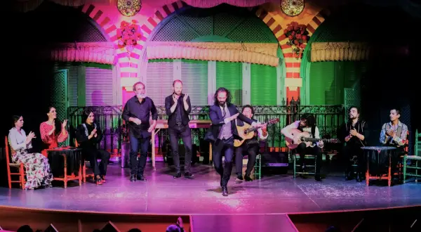 Un cantante sevillano flamenco baila en el escenario, acompañado por guitarristas y palmeros.
