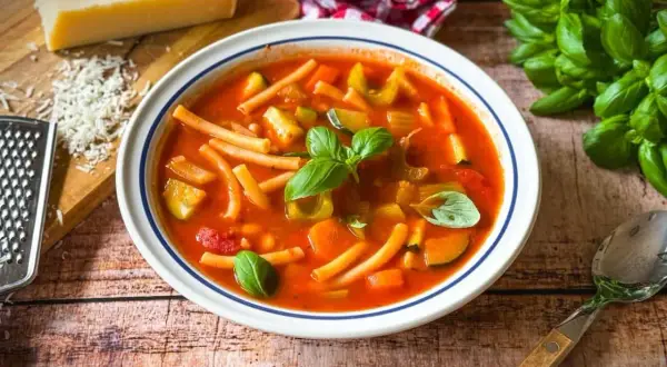 Herzhaftes rezept minestrone original mit viel Gemüse und Pasta, garniert mit frischem Basilikum. Daneben Parmesan und ein Löffel.