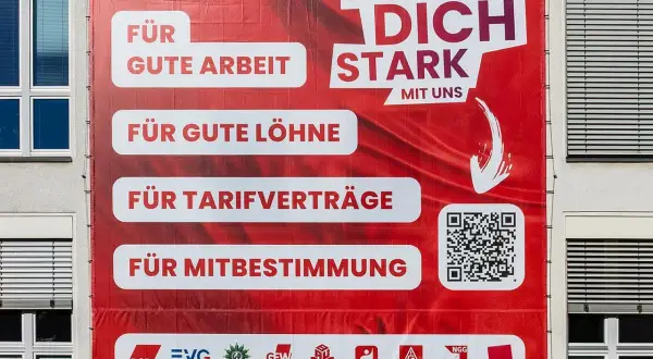 Großes rotes Banner mit der Aufschrift "Mach dich stark mit uns". Es wirbt für gute Arbeit, Löhne, Tarifverträge und Mitbestimmung. Was ist ver.di? Die Antwort steht unten.