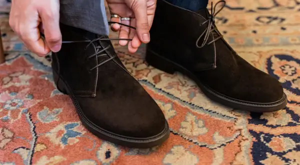 Mani che allacciano stivaletti scamosciati marroni, un dettaglio di stile per polacchi uomo eleganti su un tappeto persiano.