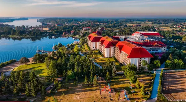 Hotel Gołębiewski w Mikołajkach, z czerwonymi dachami, otoczony zielenią i jeziorem, zaprasza na wypoczynek. Ile lat ma hotel? To miejsce z historią!