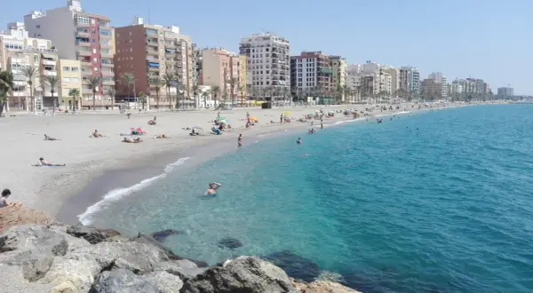 Gente disfrutando del sol y el mar en la playa del Zapillo, con edificios residenciales al fondo.
