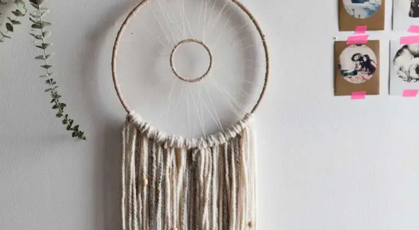 Un attrape-rêves fait main, parfait pour un tuto attrape rêve. Des fils de laine beiges et gris tombent, ornés de perles en bois.