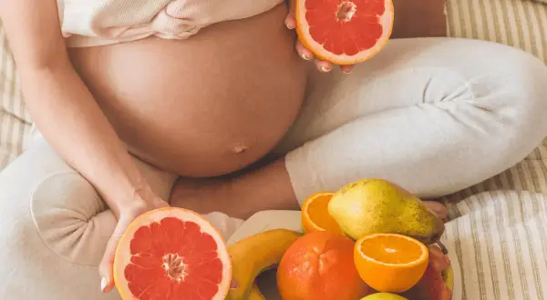 Ciężarna kobieta trzyma połówki grejpfruta, obok talerz z owocami. Czy w ciąży można jeść pomarańcze? Tak, to zdrowe!