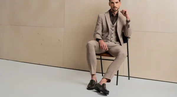 Elegancki mężczyzna w beżowym garniturze i kamizelce, siedzący na krześle. Na nogach ma stylowe loafersy, co to za buty, które dodają mu szyku.
