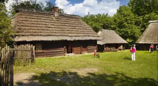 Drewniane chaty ze słomianymi dachami, trawiasty dziedziniec i ludzie.