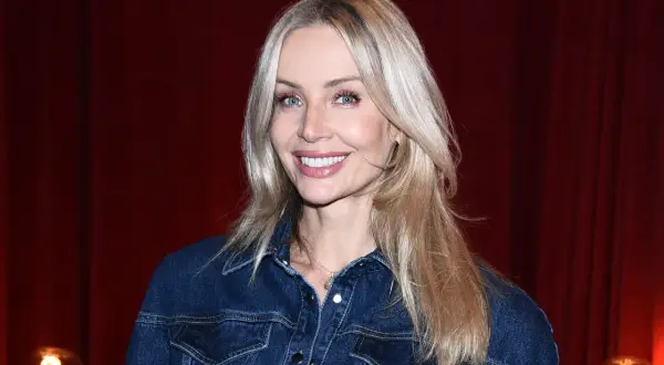 Uśmiechnięta blondynka z francuską grzywką w dżinsowej kurtce na tle czerwonej kurtyny.