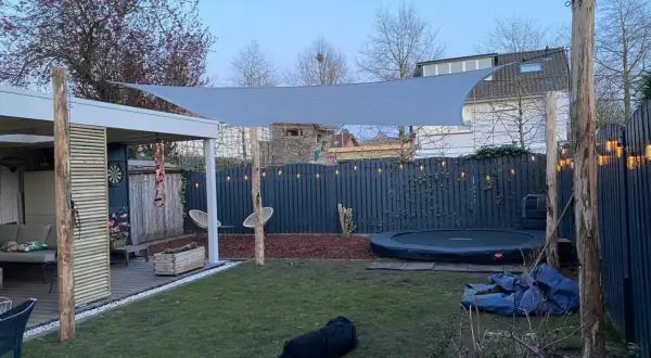 Sonnensegel befestigen Ideen: Ein graues Sonnensegel spannt sich über einen Garten mit Trampolin und Sitzbereich. Holzpfosten dienen als Befestigungspunkte.