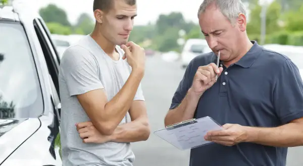 Młody mężczyzna zamyślony obok samochodu, instruktor z notatkami i długopisem przy ustach, przygotowują się do egzaminu na prawo jazdy B.
