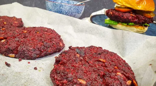 Gotowe burgery z buraka na papierze do pieczenia, jeden złożony burger z bułką i dodatkami w tle.