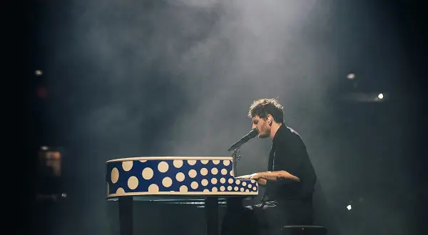 Dawid Podsiadło na koncercie, grając na pianinie w niebiesko-białe kropki, otoczony mgłą sceniczną.