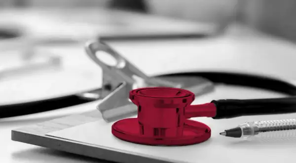 Un stéthoscope rouge vif sur un presse-papiers, prêt pour une visite médicale. Un stylo et une pince complètent la scène.