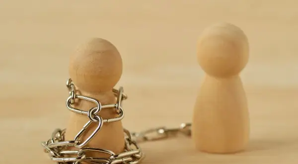 Drewniany pionek skuty łańcuchem, obok drugi, wolny. Symbolizuje to techniki manipulacji i kontroli.