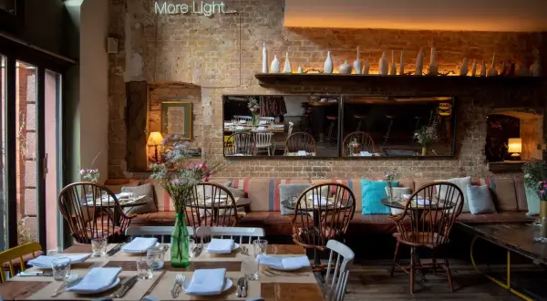 Gemütliches Restaurant mit Ziegelwänden und Tischen, gedeckt für Gäste. Eine leuchtende Neon-Schrift "More Light" hängt über dem Sitzbereich.