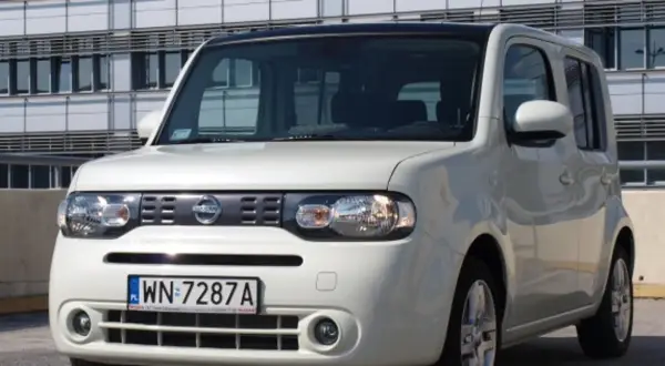 Biały Nissan Cube z polskimi tablicami rejestracyjnymi WN 7287A zaparkowany przed nowoczesnym budynkiem.