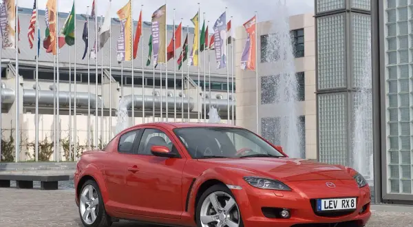 Czerwona Mazda RX-8 zaparkowana przed budynkiem z fontannami i flagami.