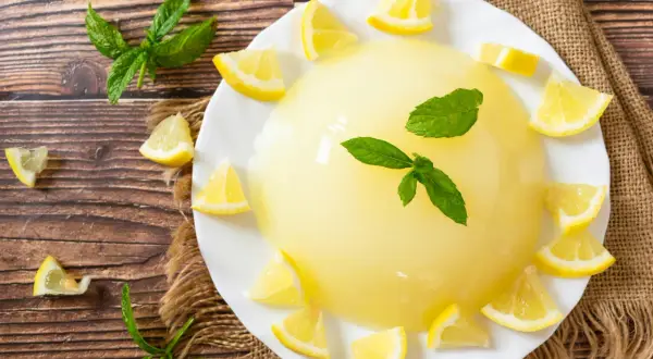 Un rinfrescante gelo di limone, guarnito con spicchi di limone e foglie di menta, su un piatto bianco, circondato da limoni e menta su un tavolo di legno.