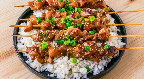 Fyra kycklingspett teriyaki ligger på en bädd av vitt ris, toppade med sesamfrön och hackad salladslök.