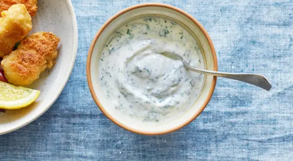 En skål med krämig kall sås till fisk, med dill och en sked. Bredvid syns friterad fisk och citronklyftor.