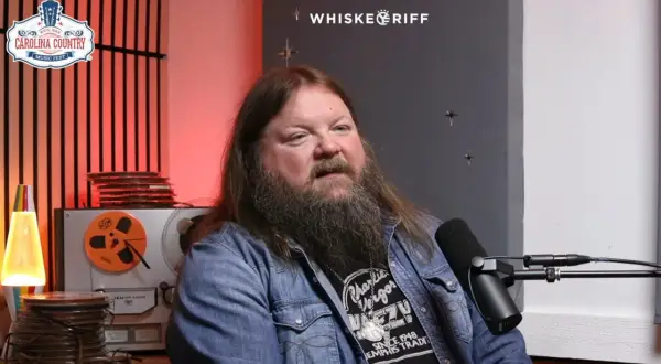 A man with a long beard and hair, wearing a denim jacket, speaks into a microphone. He's discussing Channing Wilson songs, possibly at a music festival.