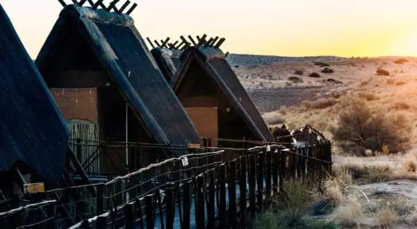 Hütten mit Strohdächern im Kgalagadi Transfrontier Park bei Sonnenuntergang. Ein Holzsteg führt zu den Unterkünften.