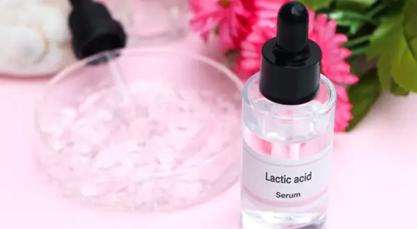 Serum z kwasem mlekowym w szklanej buteleczce z pipetą, obok różowych kwiatów.