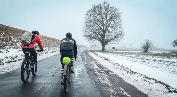 Dwaj rowerzyści na trasie Wisła 1200, w zimowej scenerii, pokonują oblodzoną drogę pod rozłożystym drzewem.