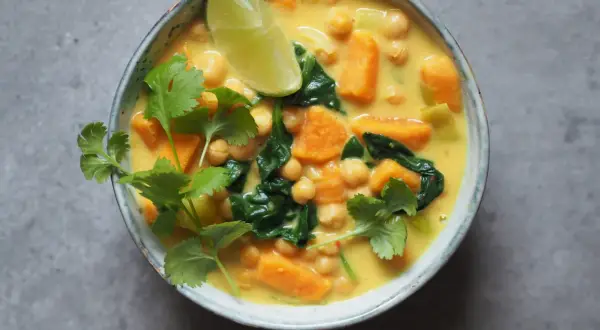 Aksamitne curry z ciecierzycy z kawałkami batata i szpinaku, ozdobione świeżą kolendrą i ćwiartką limonki.
