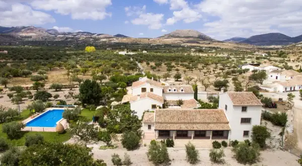 El cortijo el serbal, rodeado de olivos y montañas, con piscina azul y casas blancas de tejados terracota.