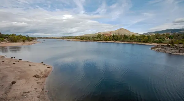 El embalse del Pontón, con aguas azules y orillas arenosas, se extiende hacia colinas verdes y un edificio de ladrillo rojo bajo un cielo nublado.