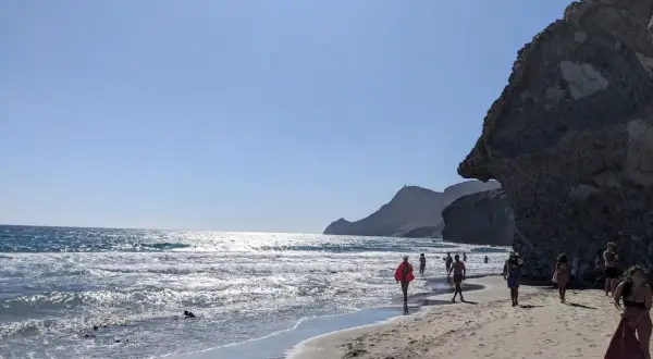Gente pasea por la **playa de Mónsul** bajo un cielo azul, con olas rompiendo en la orilla y una gran roca a la derecha.