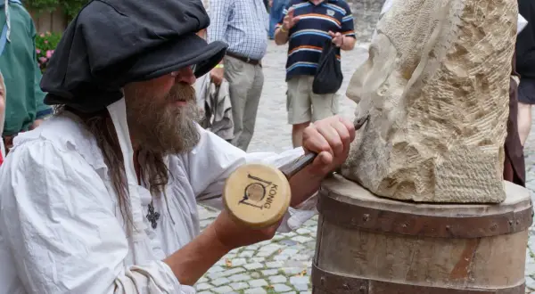 Steinmetz bei der Arbeit am Bad Wimpfen Zunftmarkt. Ein Mann mit Bart und schwarzer Kappe meißelt an einer Steinskulptur.