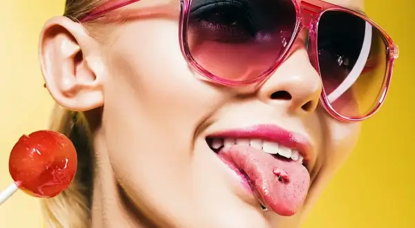 Dziewczyna w różowych okularach z lizakiem i kolczykiem w języku. Po ilu dniach można normalnie jeść po przekłuciu języka?