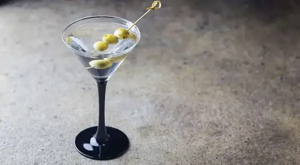 Elegancki dirty martini z trzema oliwkami na wykałaczce, stojący na czarnej podstawie.