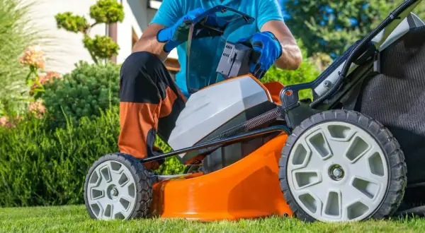 Mężczyzna wymienia baterię w pomarańczowej kosiarce akumulatorowej, przygotowując się do pracy w ogrodzie. To ważny element w rankingu kosiarek akumulatorowych.