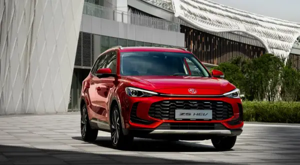 Czerwony MG ZS HEV, nowoczesny SUV, zaparkowany przed futurystycznym budynkiem.