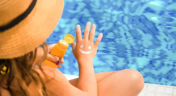 Kind mit Sonnenhut und LSF 50 Sonnencreme, die ein lachendes Gesicht auf die Hand malt.
