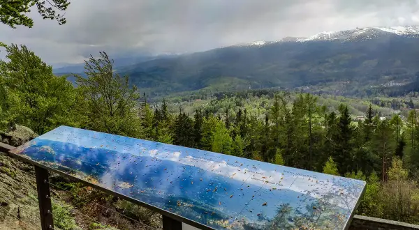 Widok z wieży widokowej Karkonosze. Na pierwszym planie tablica informacyjna z mapą panoramy, w tle góry i lasy.
