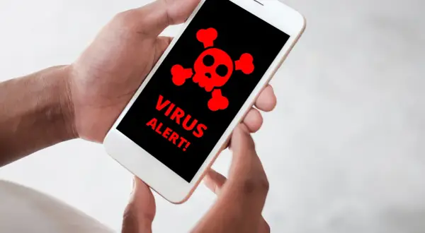 Dłonie trzymające smartfon z czerwonym symbolem czaszki i napisami "VIRUS ALERT!".