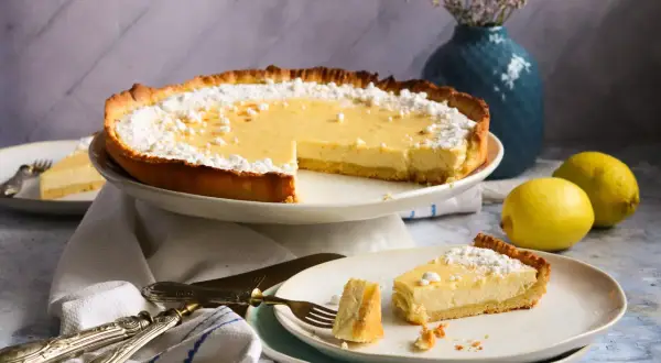 Eine köstliche Tarte au Citron, bestäubt mit Puderzucker, steht bereit. Ein Stück wurde bereits auf einem Teller serviert, daneben liegen frische Zitronen.